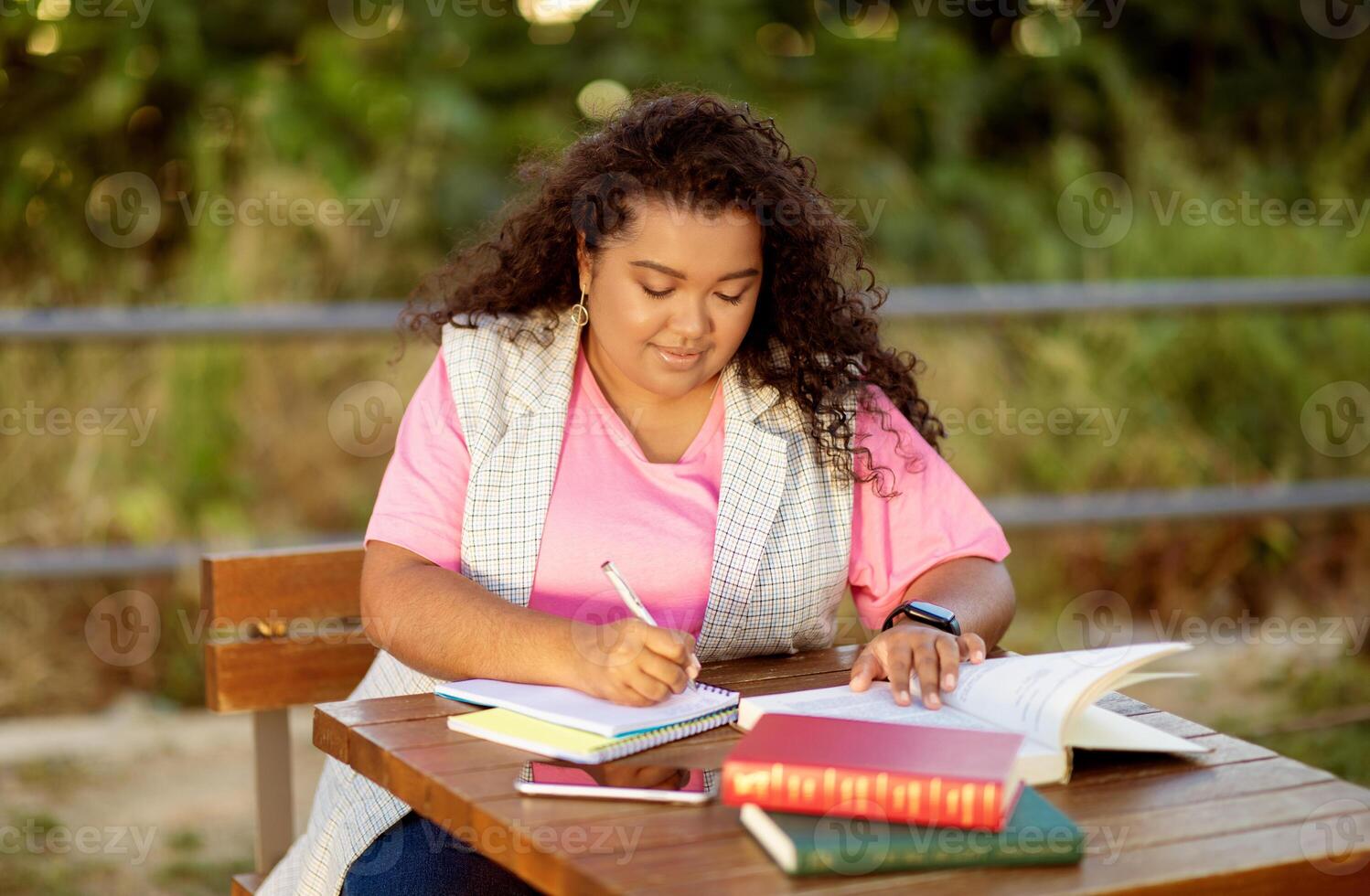 A student engages in studying at a wooden table outside, surrounded by nature. She writes in her notebook while several books lie open, showcasing her dedication to learning. photo