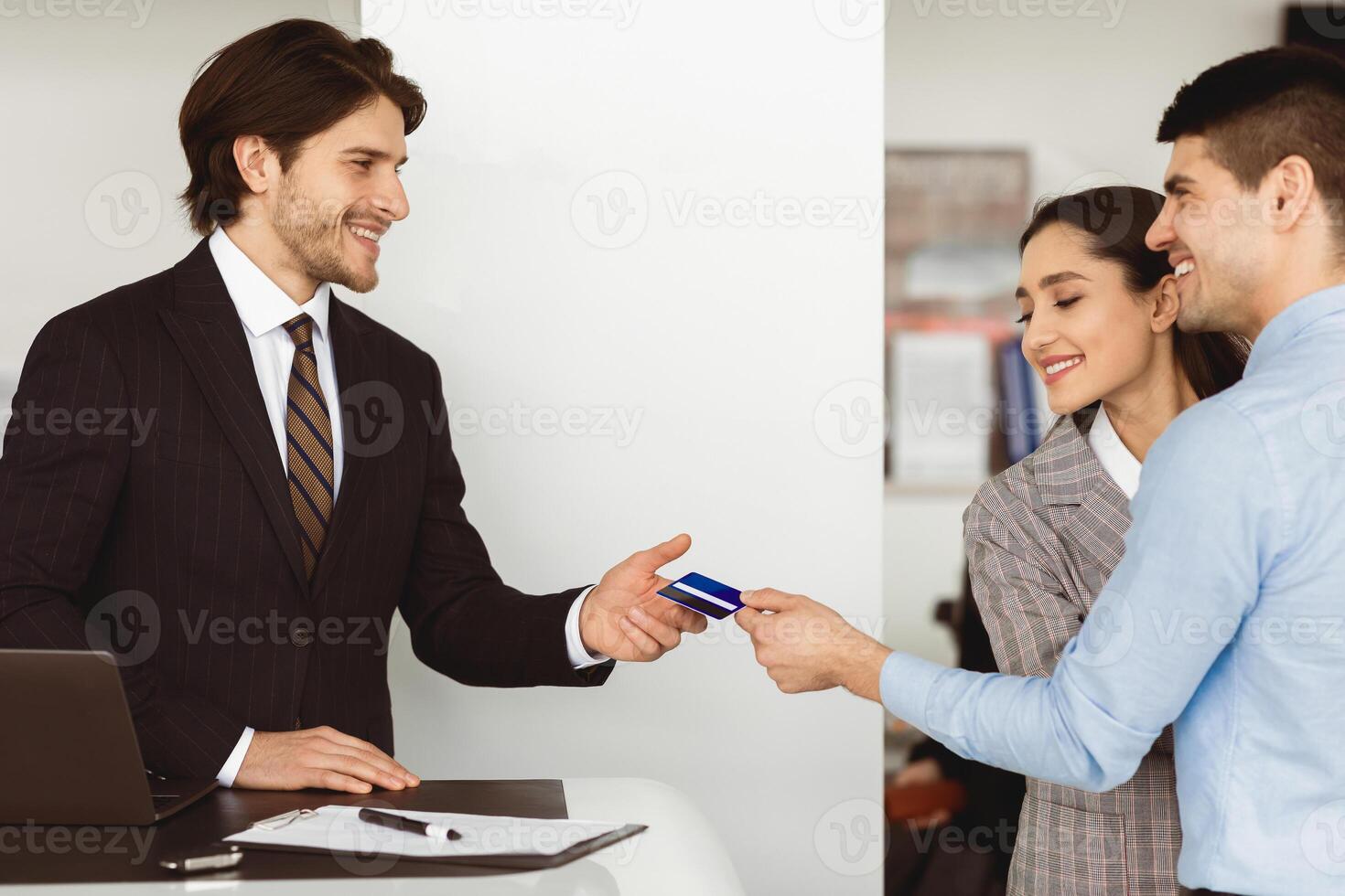 Happy Spouses Buying Car Giving Seller Credit Card Standing In Auto Dealership Office. Empty Space. Side View photo