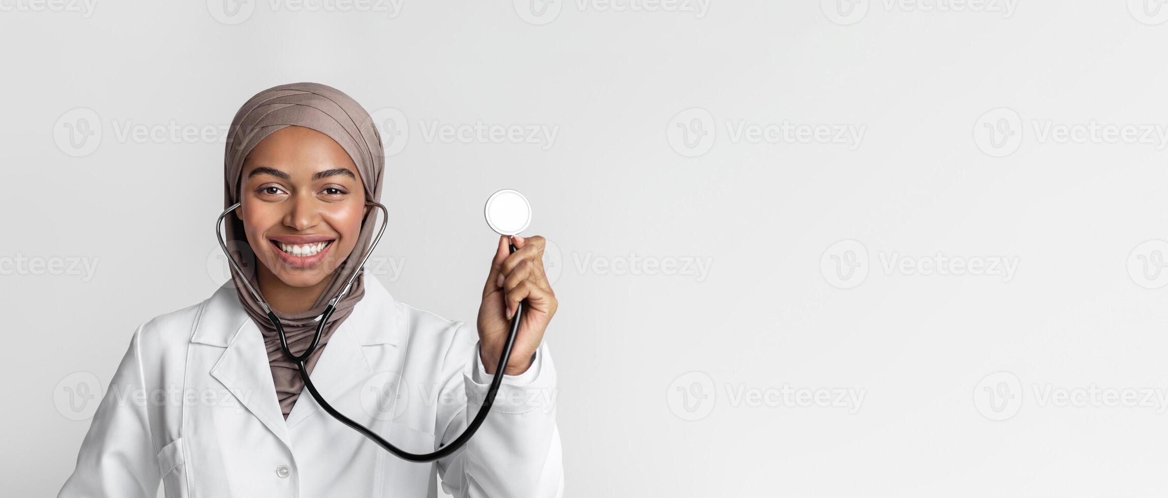 Portraif of young black muslim female doctor in hijab with stethoscope, posing over light background and smiling at camera, free space photo