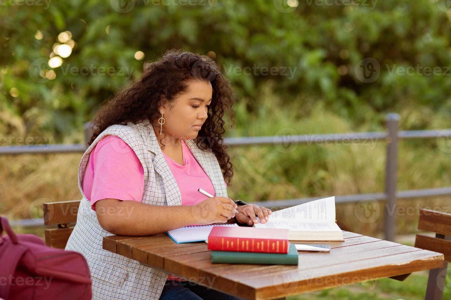 A student engaged in studying while seated at a wooden table outdoors. The individual writes in a notebook, with books beside them, immersed in their studies on a sunny day. photo