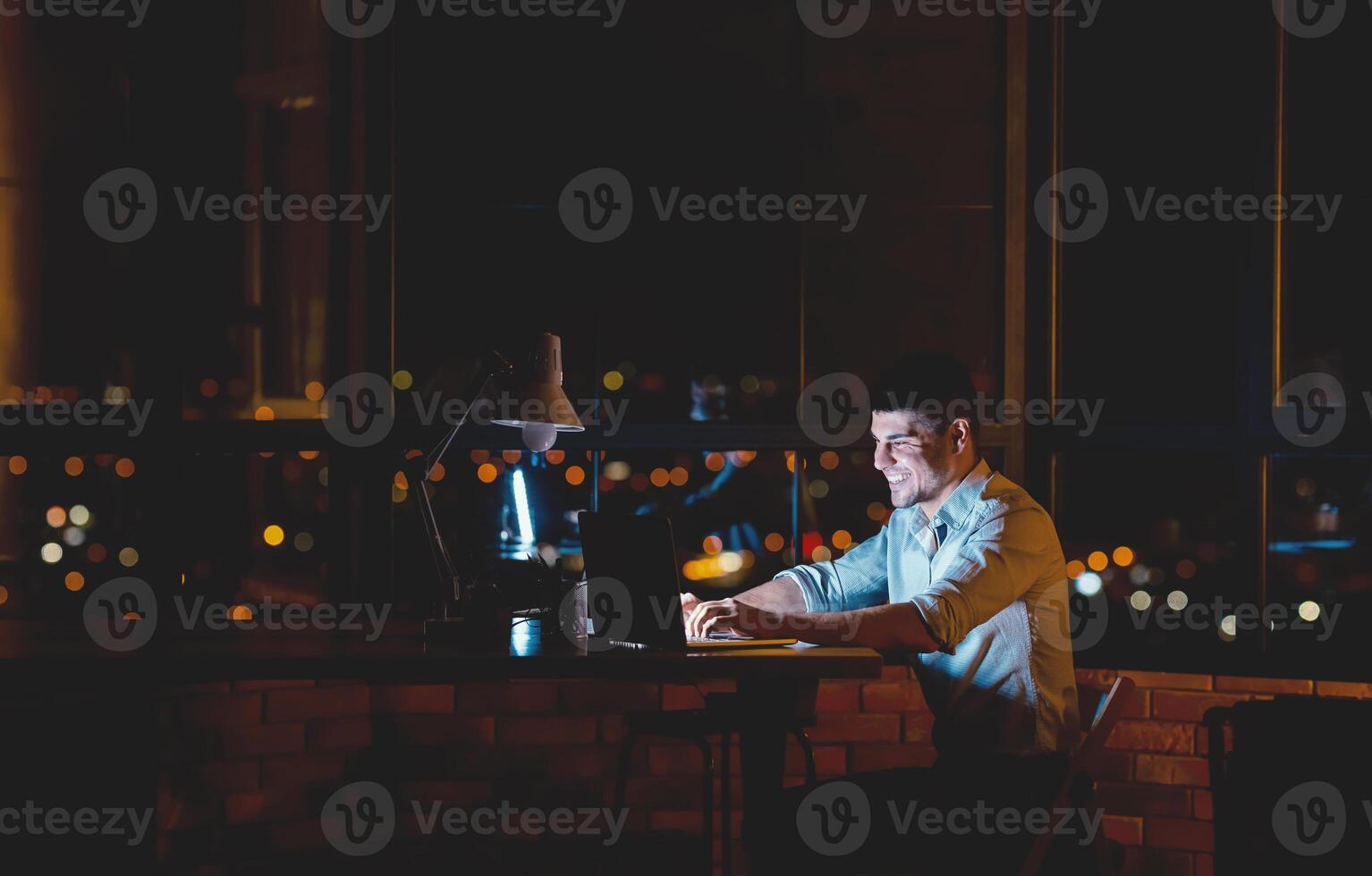 Joyful Man Working On Laptop Computer Browsing Internet Sitting Late At Night In Modern Office. Empty Space For Text photo
