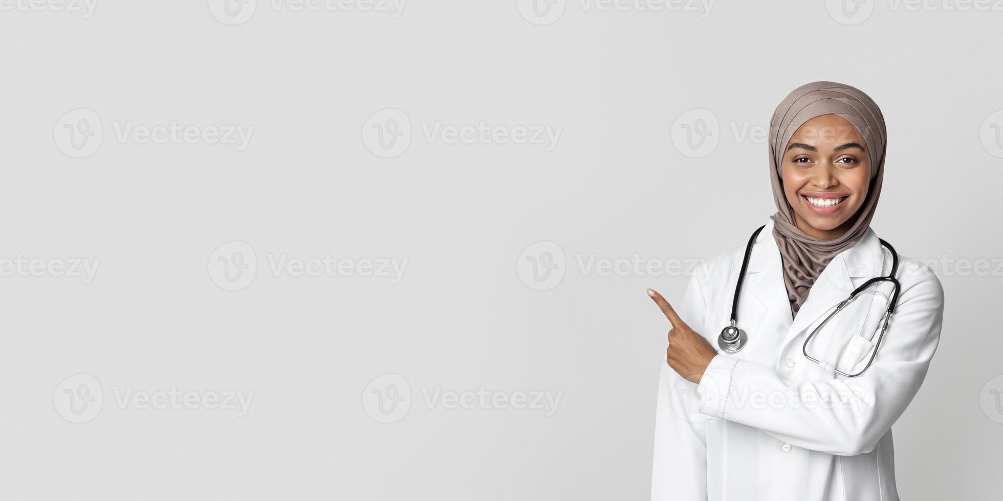 Positive black female therapist in hijab and white coat pointing aside at copy space on light background, indicating free place for your ad, panorama photo