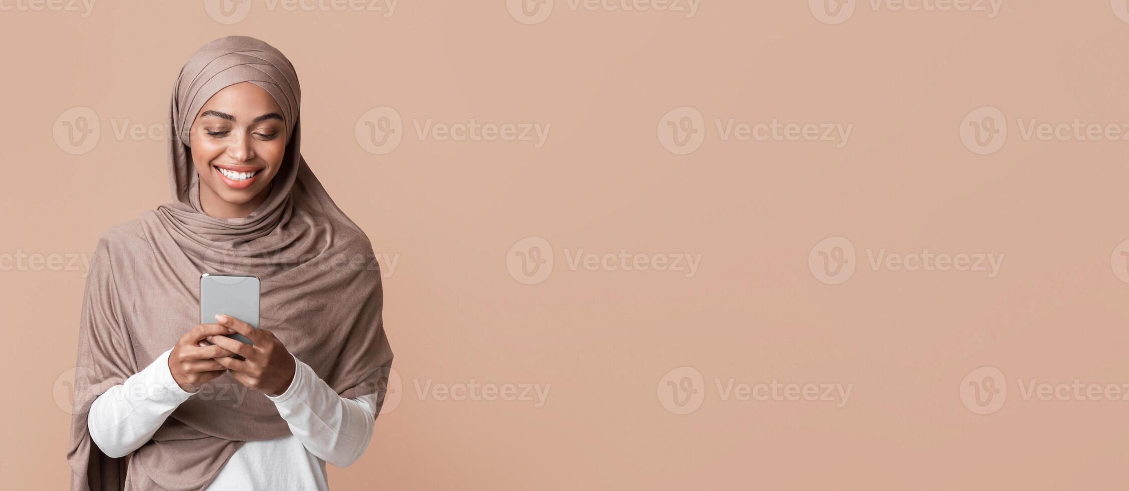 Smiling afro muslim girl in hijab using smartphone, texting or downloading new app, standing over yellow background with empty space photo