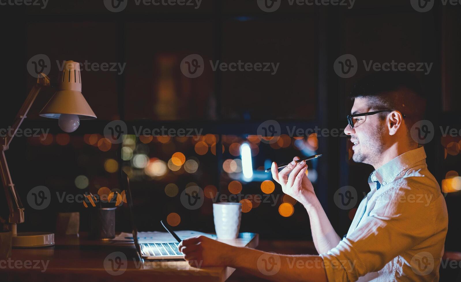Business Lifestyle. Man Using Smartphone Voice Assistant Working On Laptop In Modern Office At Night. Copy Space photo