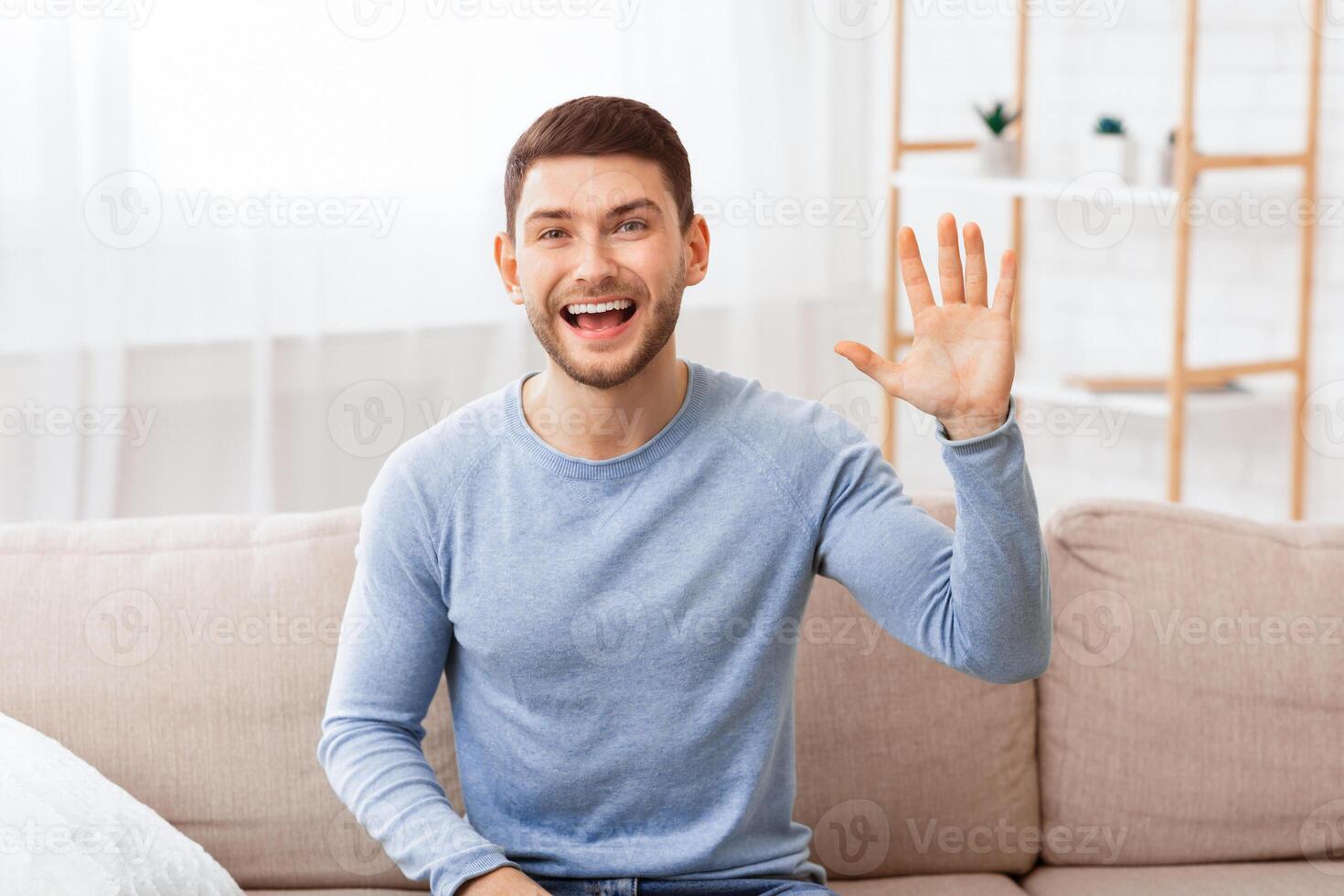 Hello. Cheerful Guy Waving Hand Smiling Looking At Camera Sitting On Couch Indoor. photo