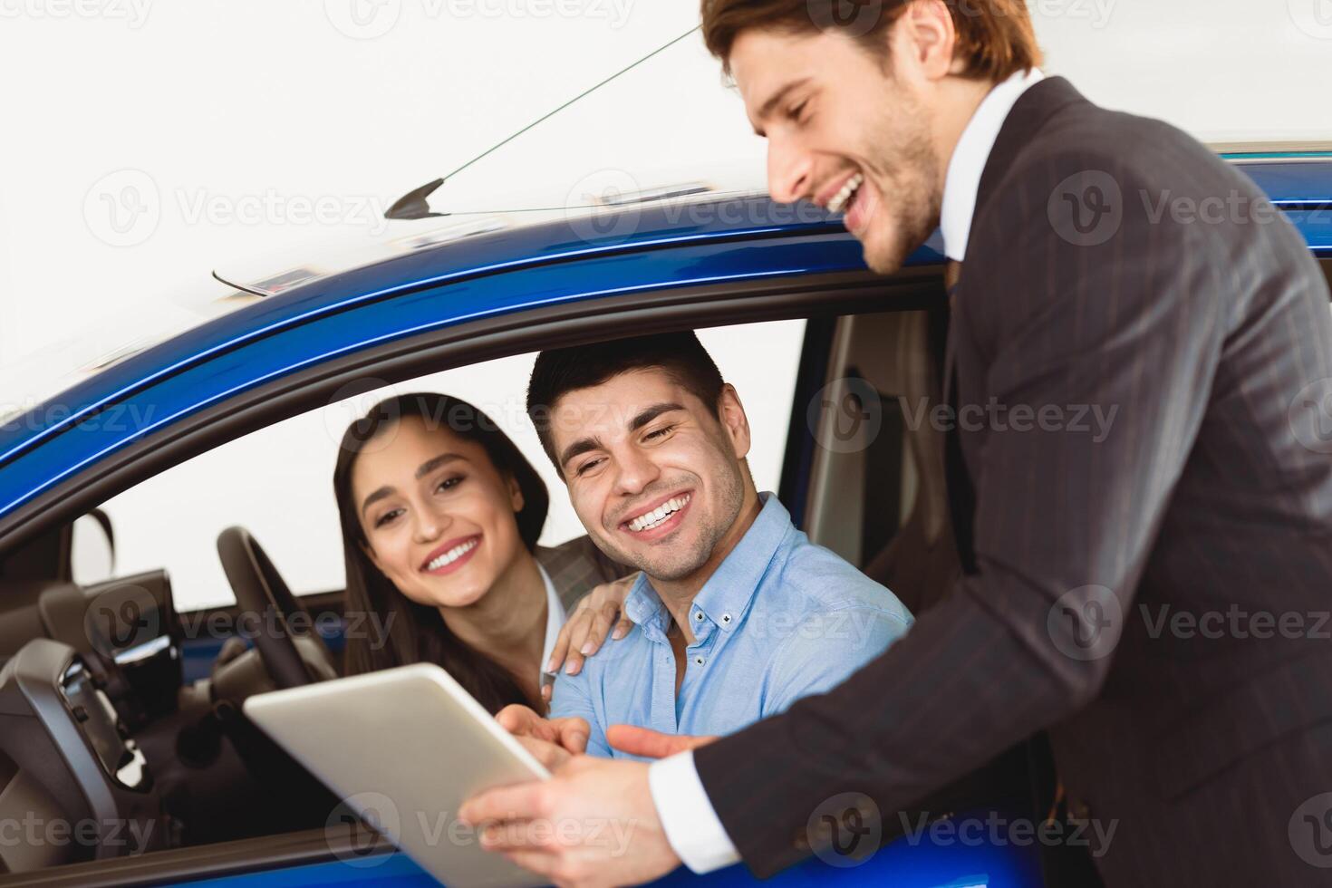 Happy Couple Testing New Car, Talking With Salesman Who Showing Them Agreement On Digital Tablet photo