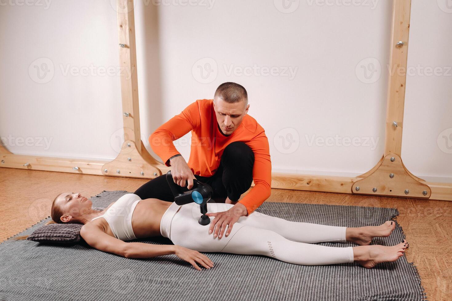 A male masseur massages a girl's body with a percussion massager to prepare for a stretching simulator photo