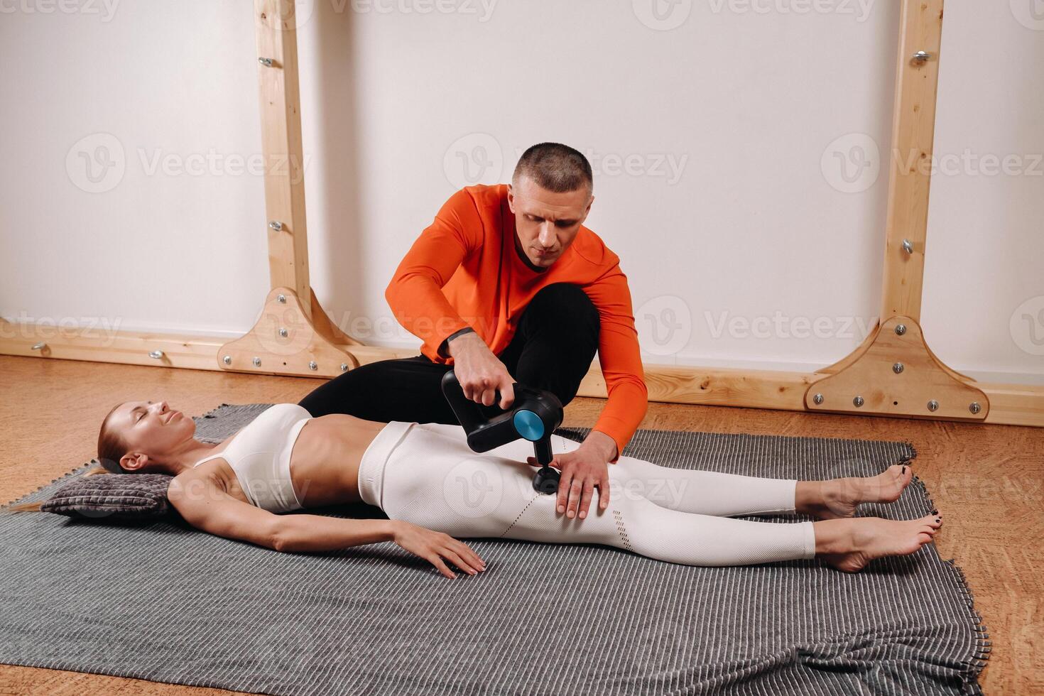 A male masseur massages a girl's body with a percussion massager to prepare for a stretching simulator photo