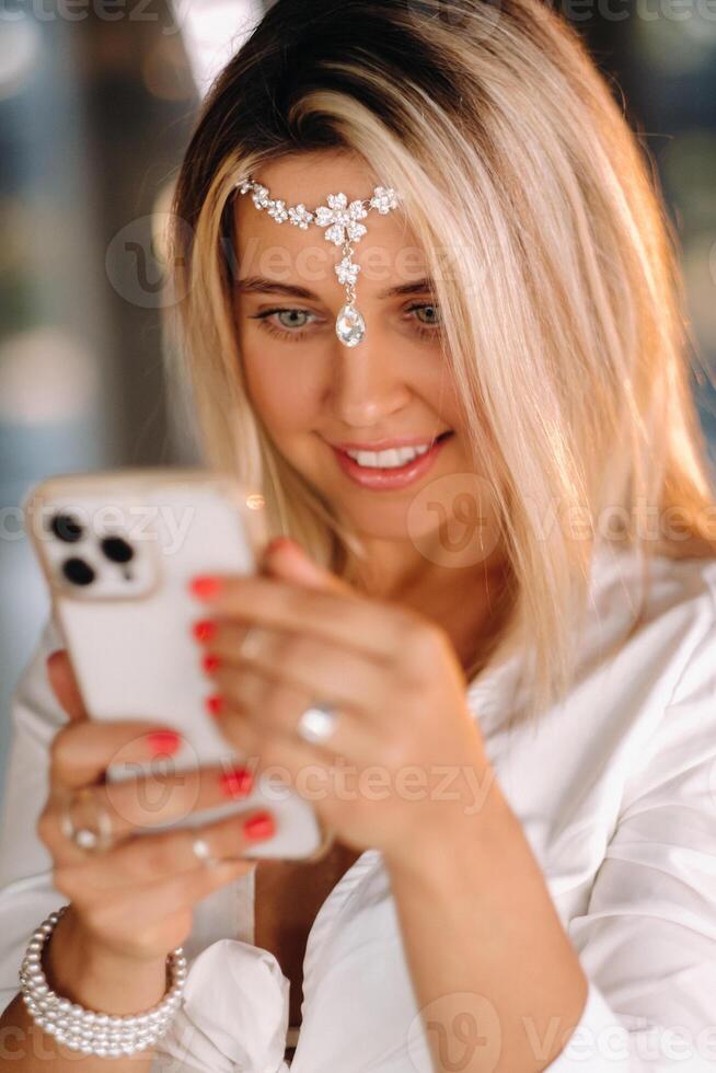 A positive young woman in a white dress and an ornament on her head was smiling, holding a phone in her hands and looking into it photo