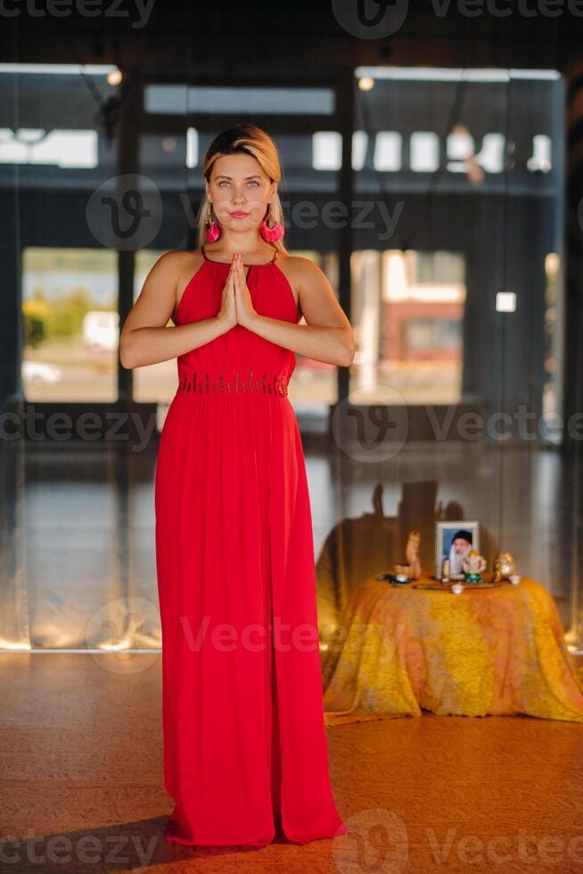 Portrait of a smiling girl in a red dress stands in the interior with her hands clasped in front of her photo