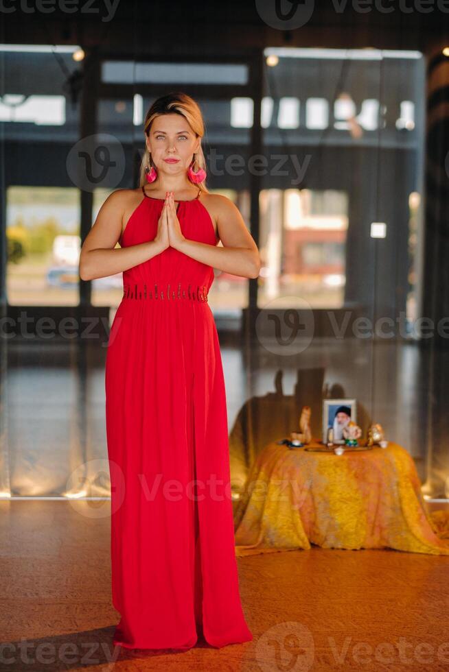 Portrait of a smiling girl in a red dress stands in the interior with her hands clasped in front of her photo