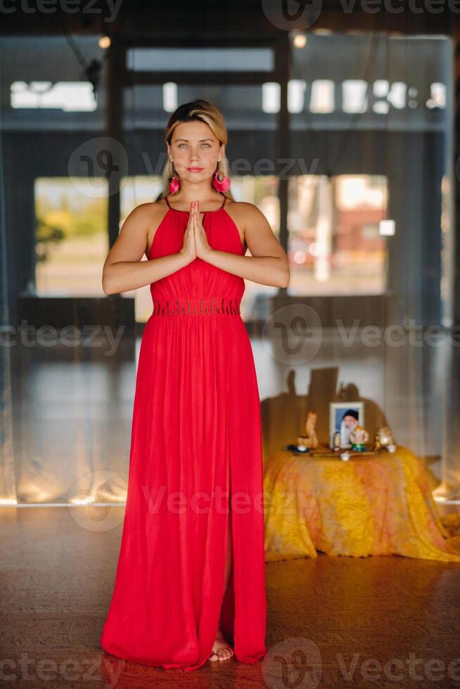 Portrait of a smiling girl in a red dress stands in the interior with her hands clasped in front of her photo