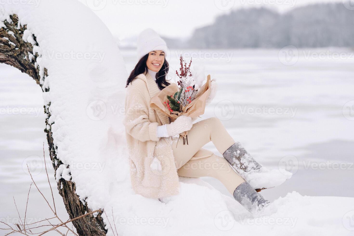 A girl in a beige cardigan with winter flowers is sitting in nature in a snowy park. Winter weather photo