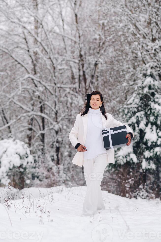 A stylish woman with a white suit with a New Year's gift in her hands in a winter forest. A girl in nature in a snowy forest with a gift box photo