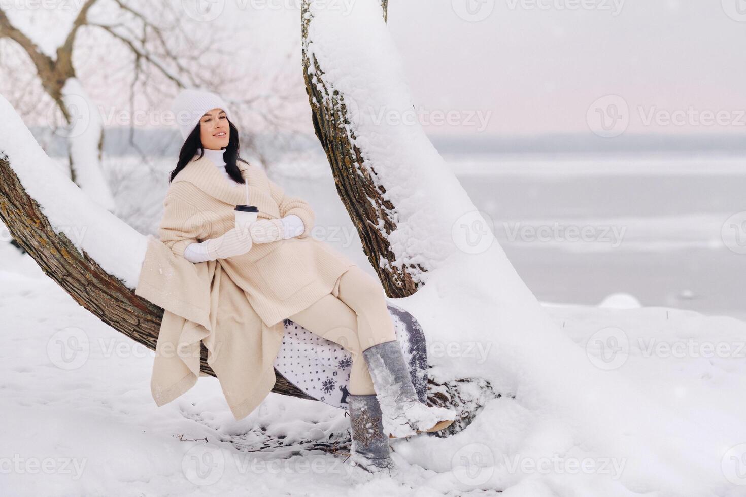 A beautiful girl with a beige cardigan and a white hat enjoying drinking tea in a snowy winter forest near a lake photo