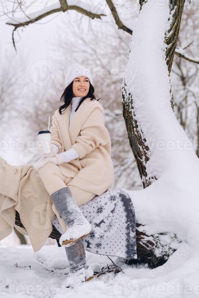 A beautiful girl with a beige cardigan and a white hat enjoying drinking tea in a snowy winter forest near a lake photo