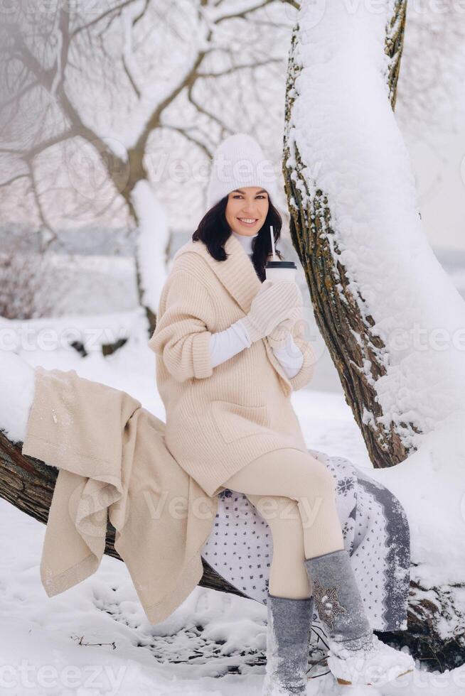 A beautiful girl with a beige cardigan and a white hat enjoying drinking tea in a snowy winter forest near a lake photo