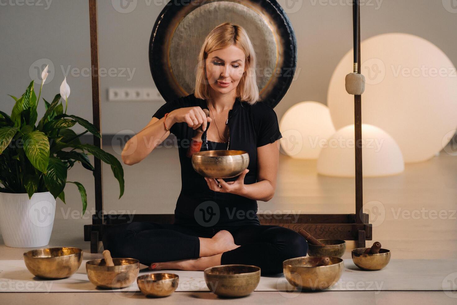 A woman plays a Tibetan singing bowl while sitting on a yoga mat against the background of a gong photo