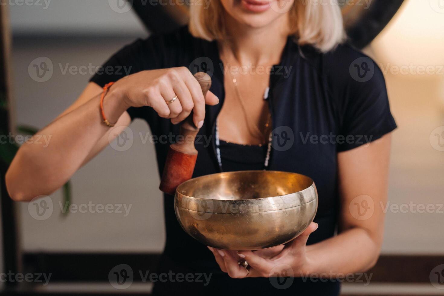 A woman plays a Tibetan singing bowl while sitting on a yoga mat against the background of a gong photo