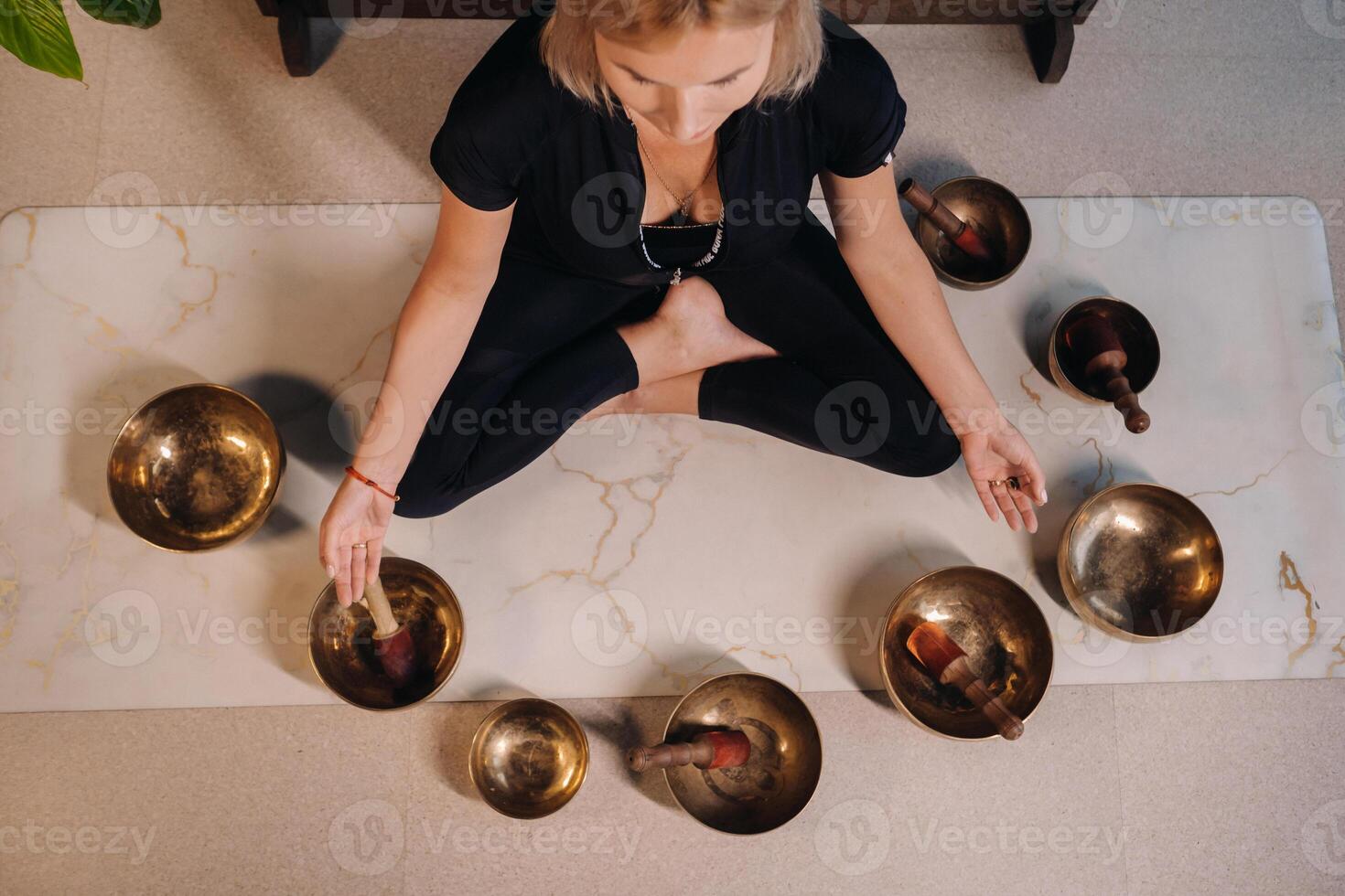 A woman sits in a lotus position next to Tibetan bowls, sitting on a yoga mat against the background of a gong photo