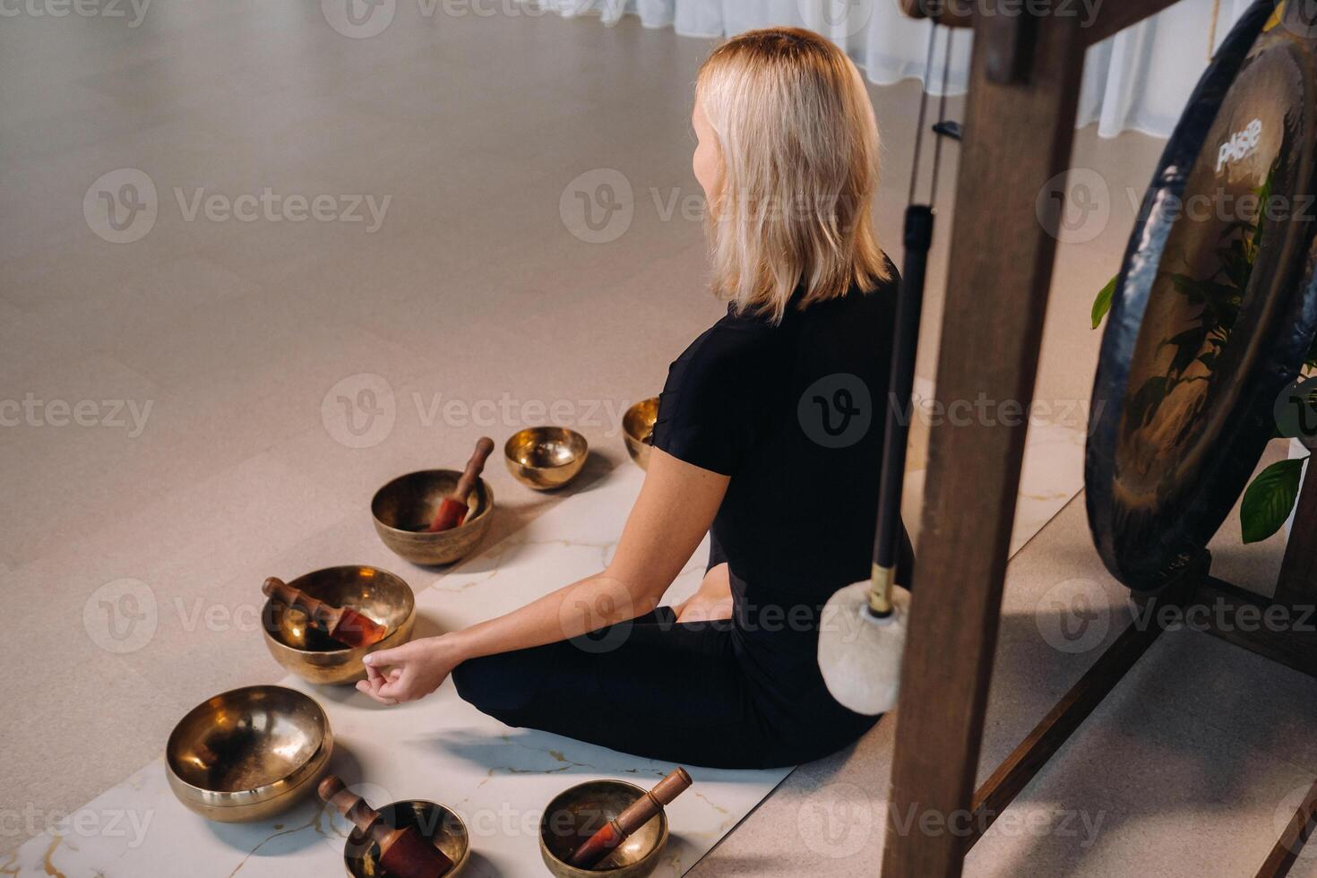 A woman sits in a lotus position next to Tibetan bowls, sitting on a yoga mat against the background of a gong photo