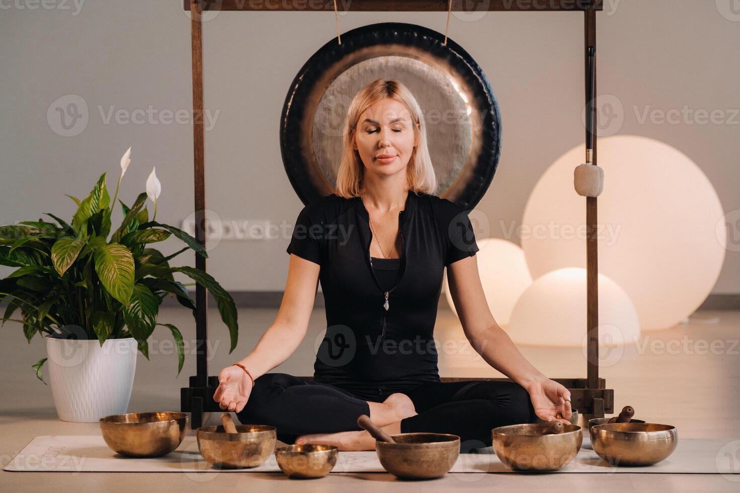 A woman sits in a lotus position next to Tibetan bowls, sitting on a yoga mat against the background of a gong photo