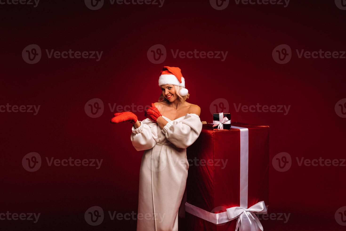 portrait of a smiling girl in a white dress and Santa hat with a huge Christmas gift on a red background photo
