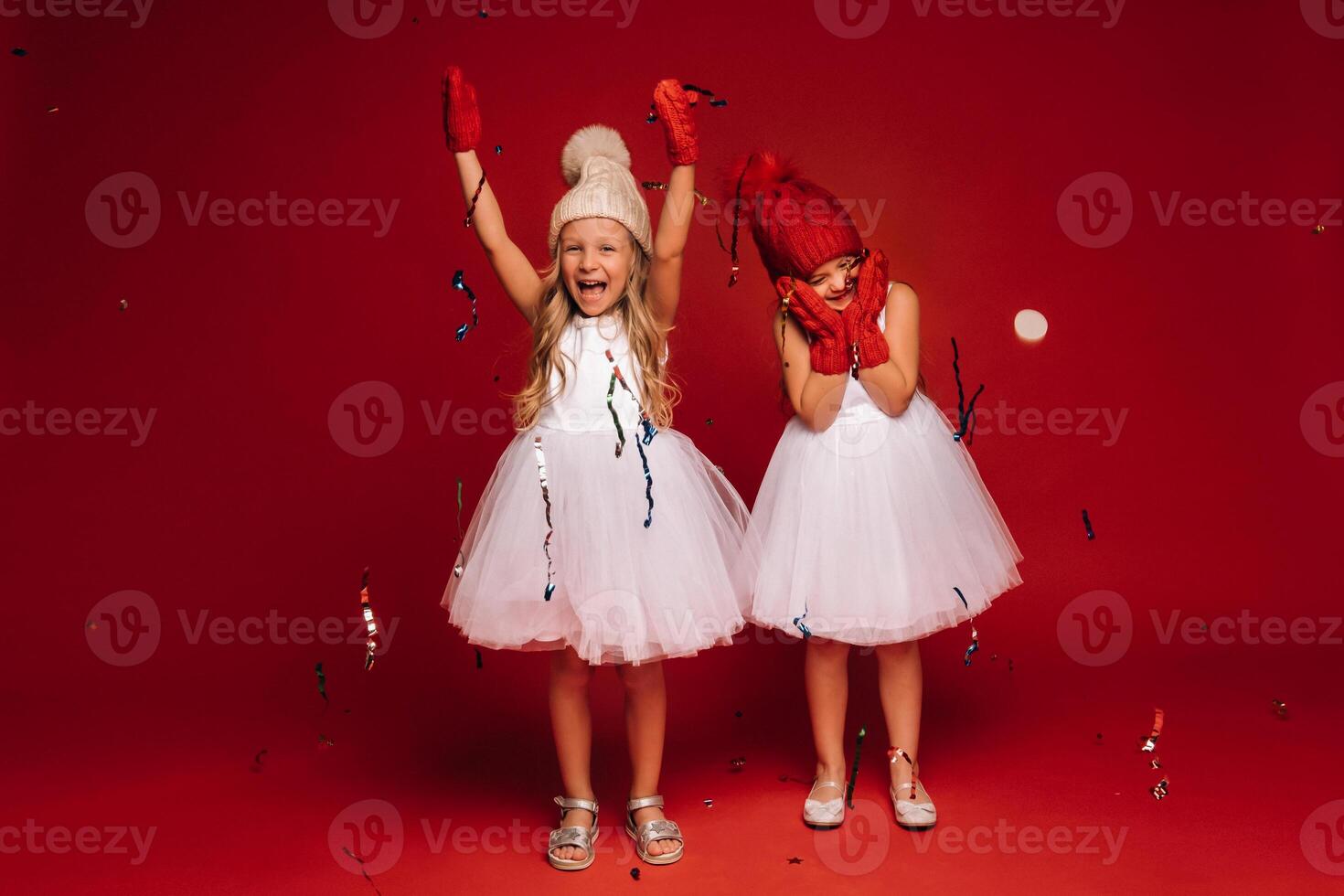 two little girls in white dresses hats and mittens sprinkle confetti on a red background photo