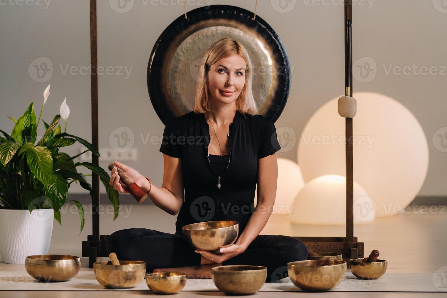 A woman plays a Tibetan singing bowl while sitting on a yoga mat against the background of a gong photo