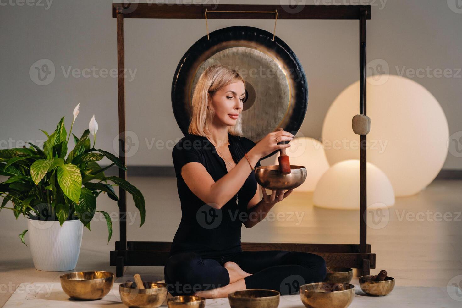 A woman plays a Tibetan singing bowl while sitting on a yoga mat against the background of a gong photo