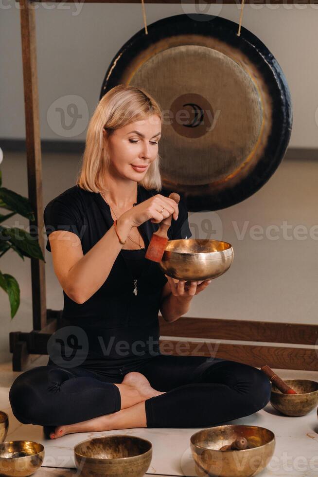 A woman plays a Tibetan singing bowl while sitting on a yoga mat against the background of a gong photo