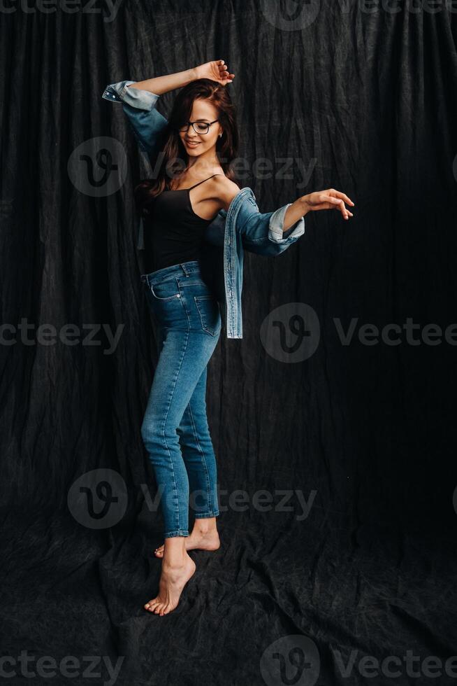 a brunette with long hair and glasses poses in the studio standing on a black background photo
