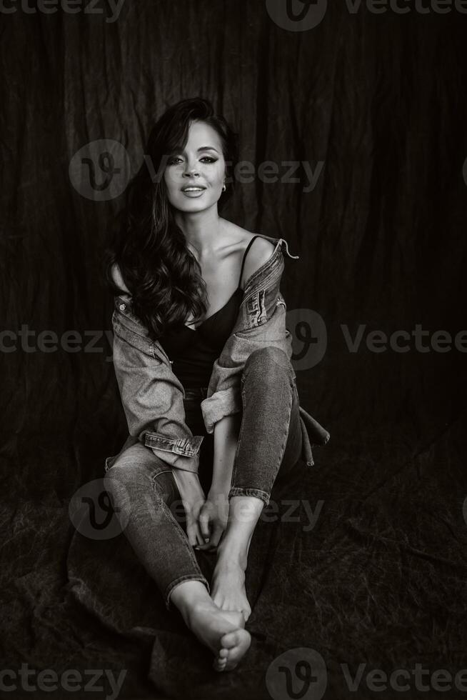 a brunette with long hair poses in the studio sitting on the floor on a black background.Black and white photo