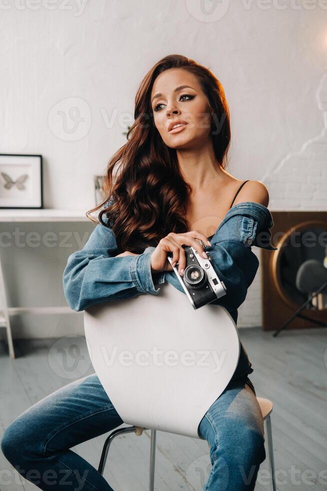 a brunette with long hair poses in a studio sitting on a chair with a camera in her hands photo