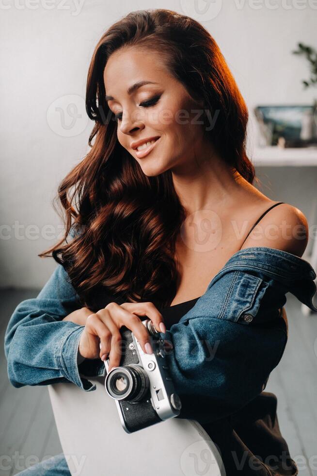 a brunette with long hair poses in a studio sitting on a chair with a camera in her hands photo