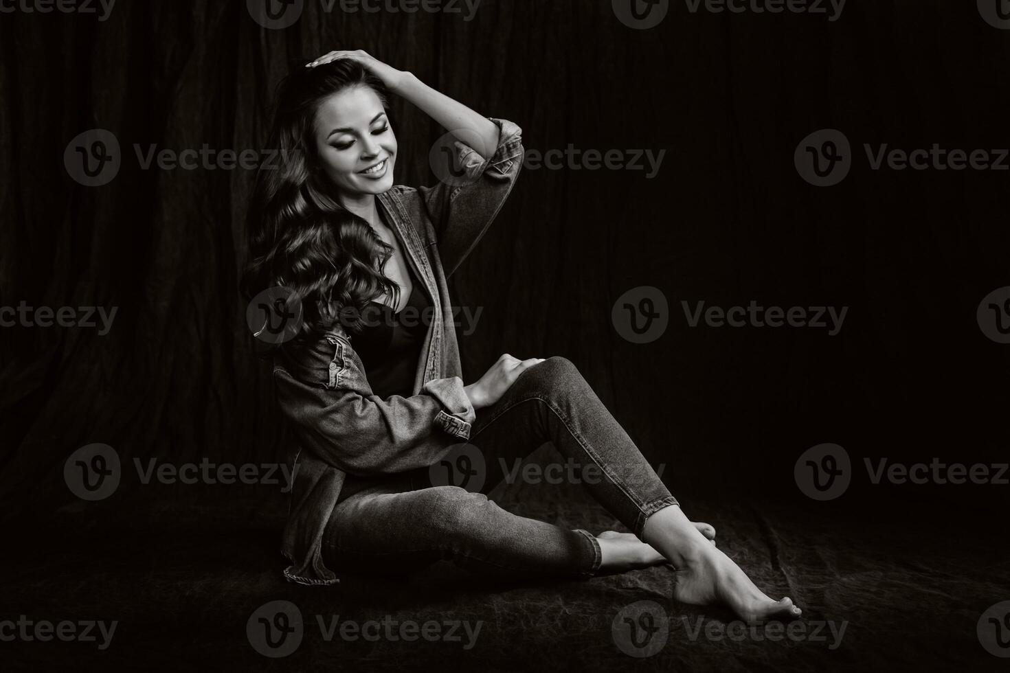 a brunette with long hair poses in the studio sitting on the floor on a black background.Black and white photo