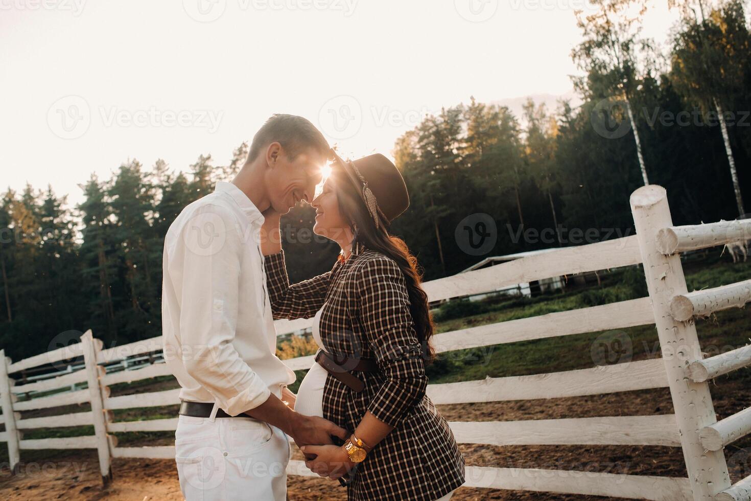 a pregnant girl in a hat and her husband in white clothes stand next to a horse corral at sunset.a stylish couple is waiting for a child in nature photo