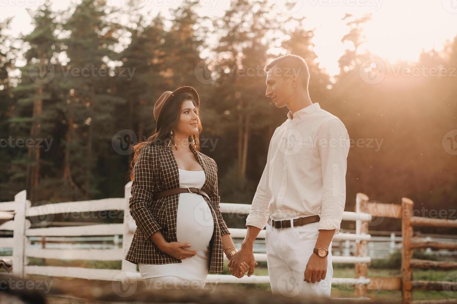 a pregnant girl in a hat and her husband in white clothes stand next to a horse corral at sunset.a stylish couple is waiting for a child in nature photo