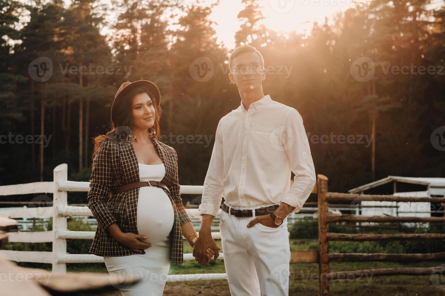 a pregnant girl in a hat and her husband in white clothes stand next to a horse corral at sunset.a stylish couple is waiting for a child in nature photo