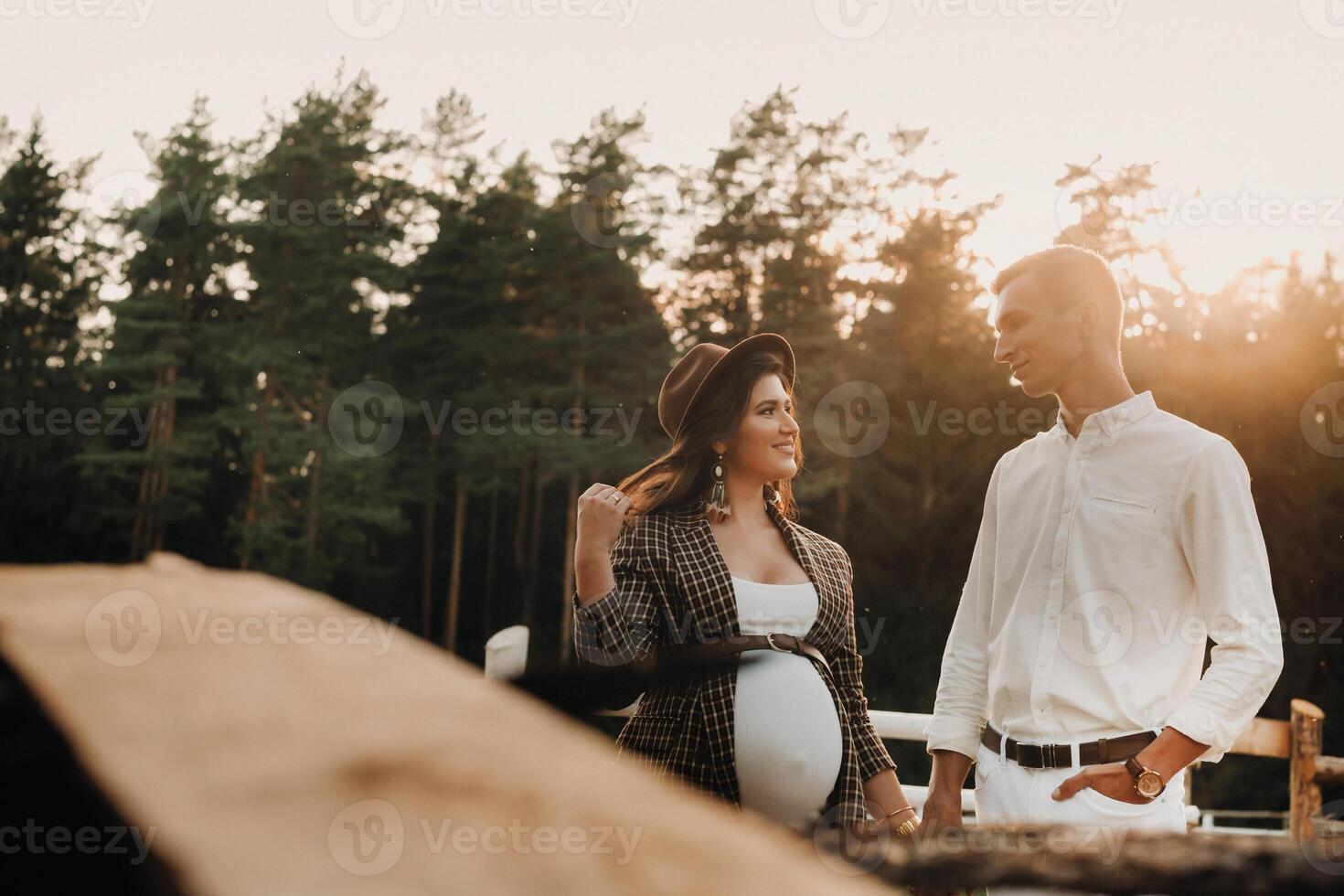 a pregnant girl in a hat and her husband in white clothes stand next to a horse corral at sunset.a stylish couple is waiting for a child in nature photo
