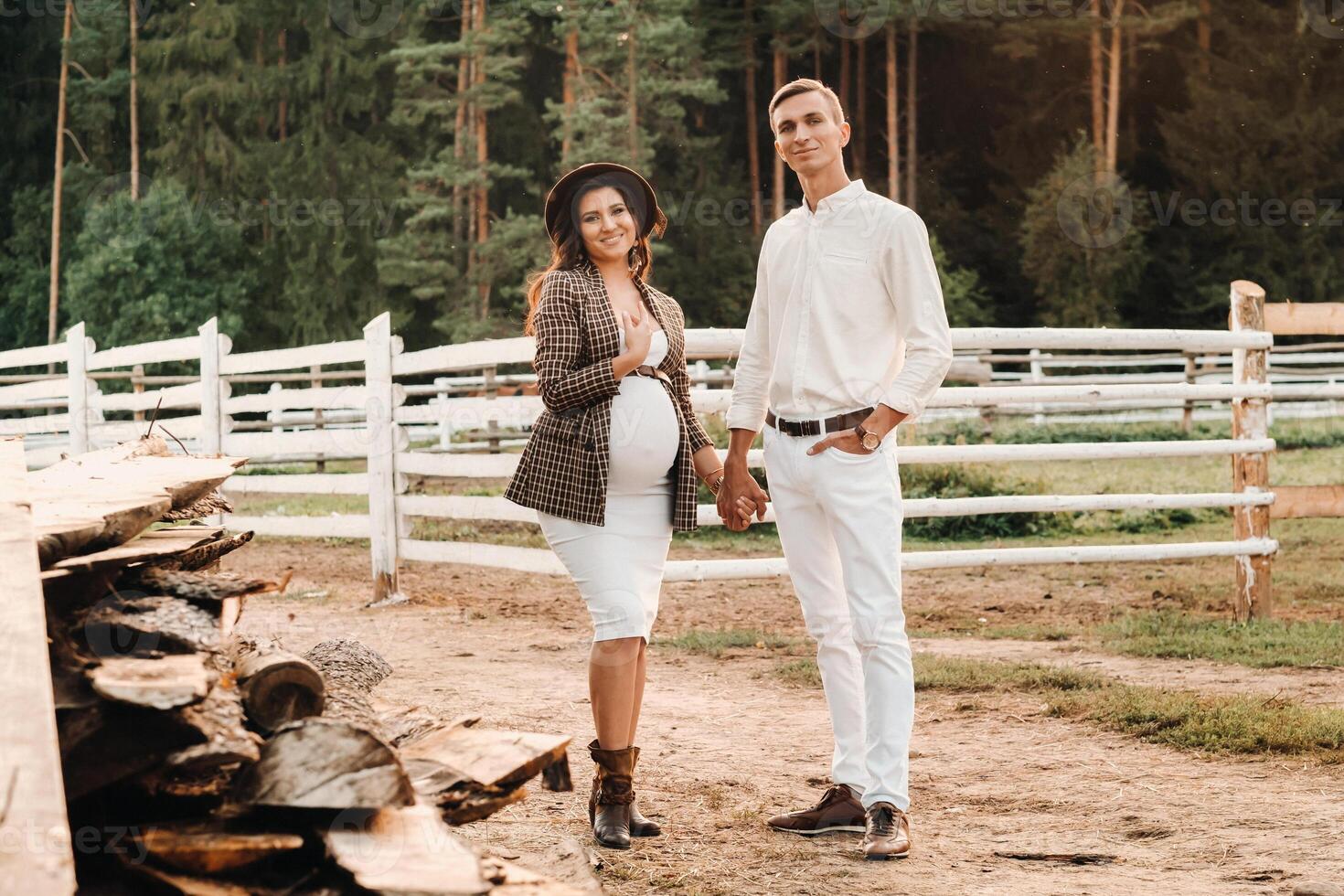 a pregnant girl in a hat and her husband in white clothes stand next to a horse corral at sunset.a stylish couple is waiting for a child in nature photo