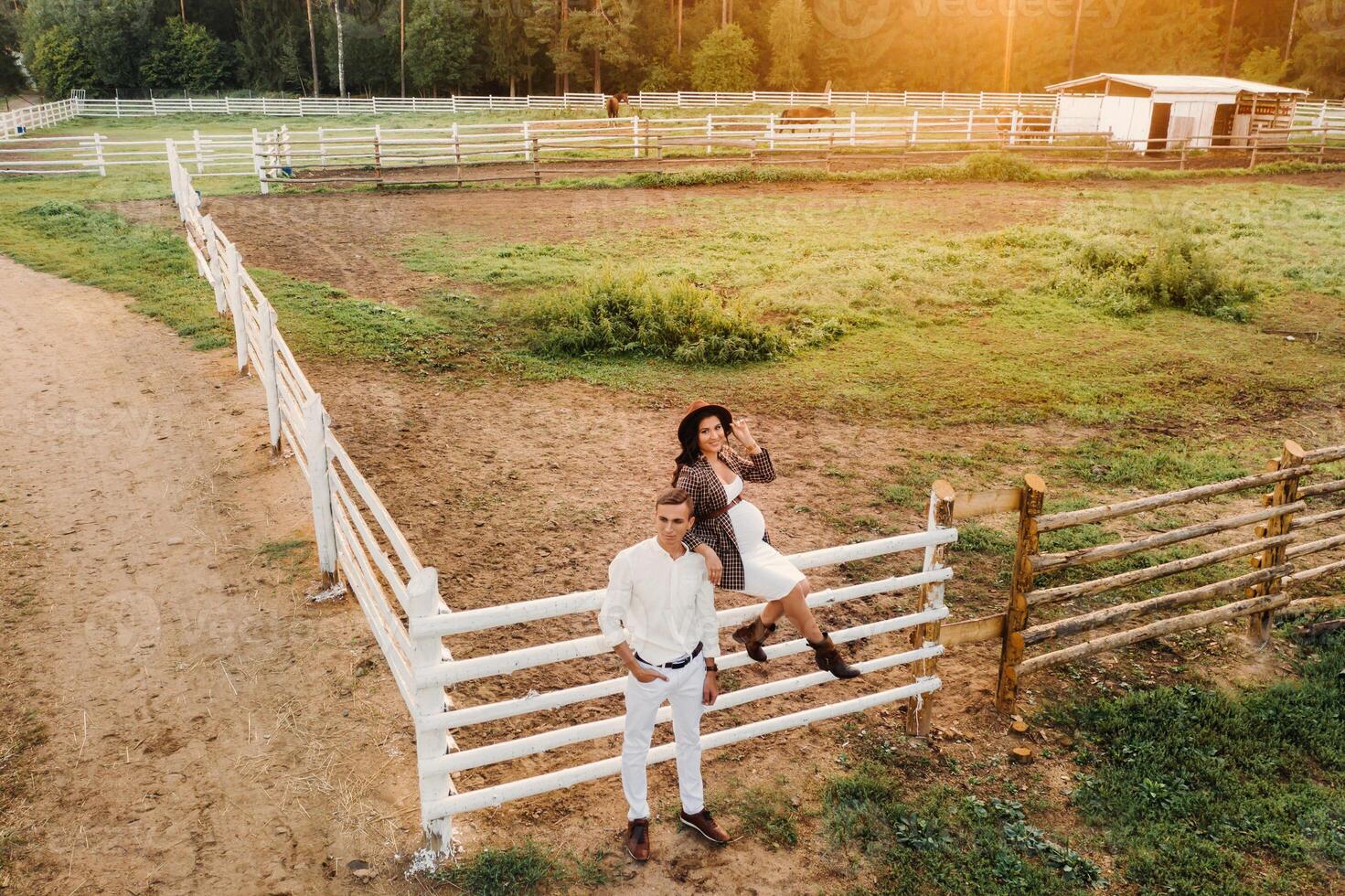 a pregnant girl in a hat and her husband in white clothes stand next to a horse corral at sunset.a stylish couple is waiting for a child in nature photo