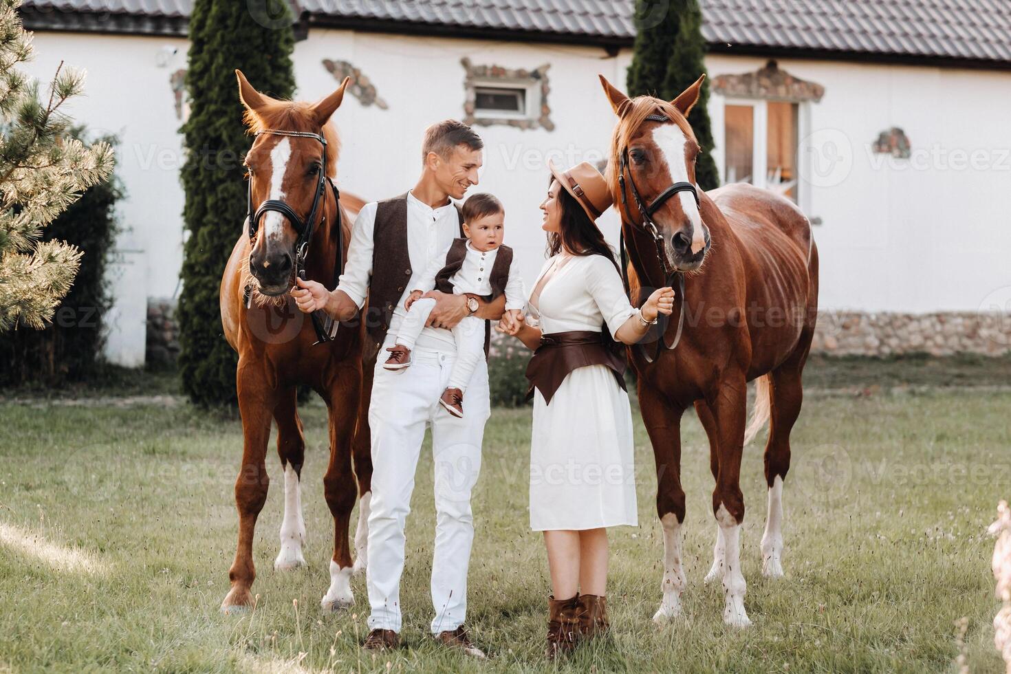 A family in white clothes with their son stand near two beautiful horses in nature. A stylish couple with a child are photographed with horses photo