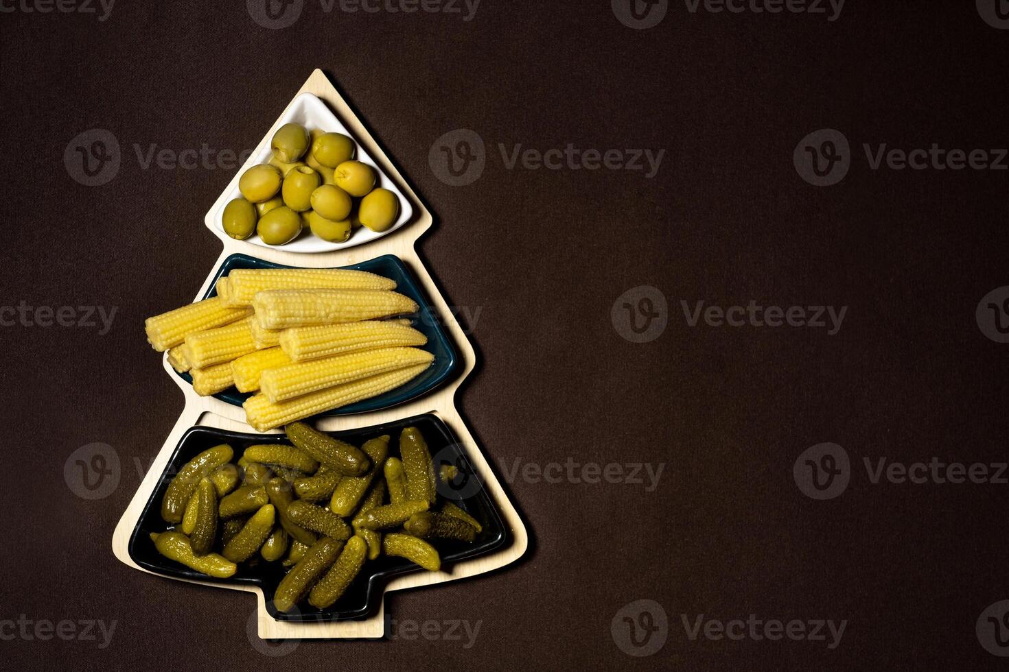 Dish in the form of a Christmas tree with pickled vegetables on a brown background.A plate of cucumbers, corn and olives at Christmas. photo