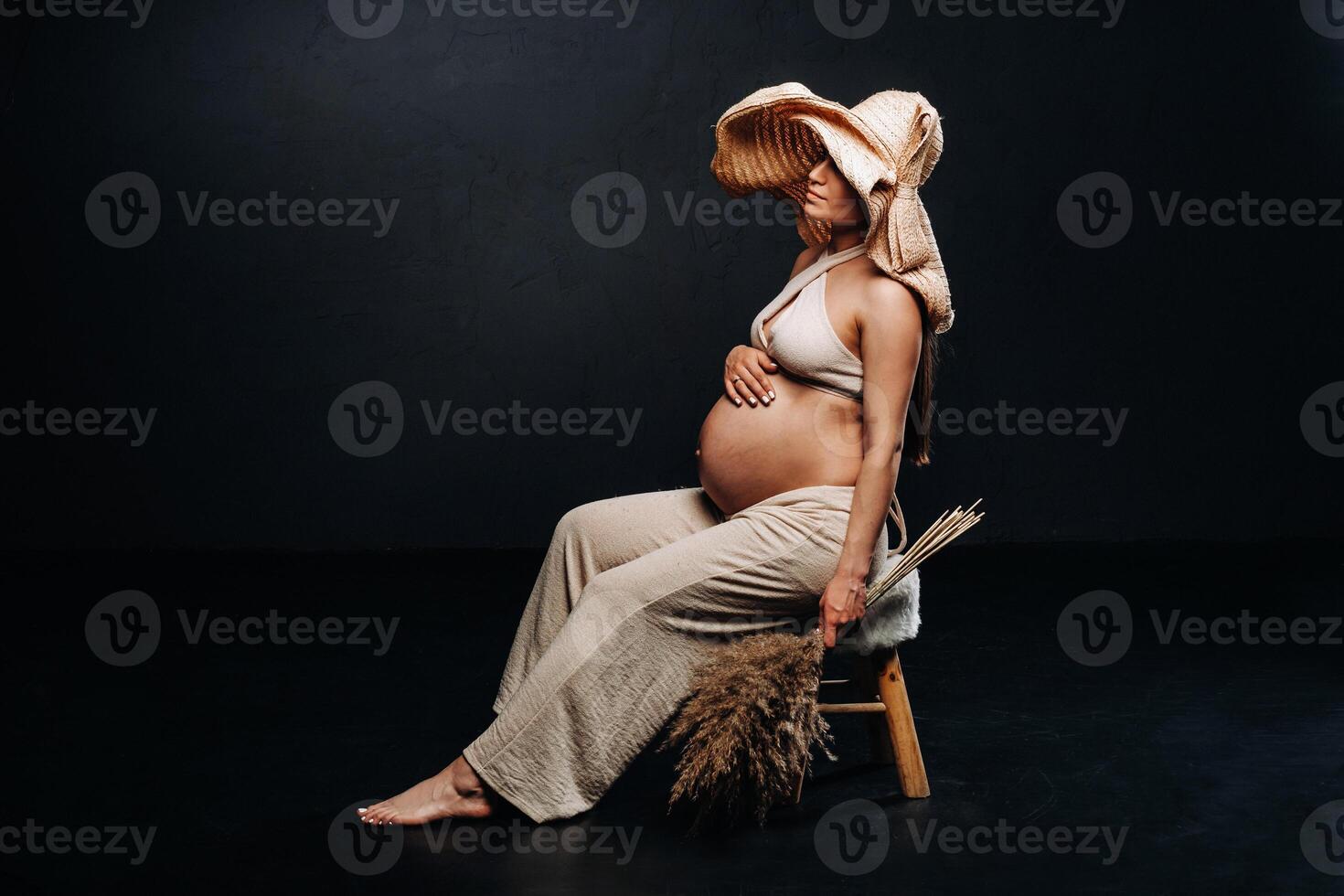 pregnant woman in a straw hat with beige clothes with a bouquet in her hands in the studio on a black background photo