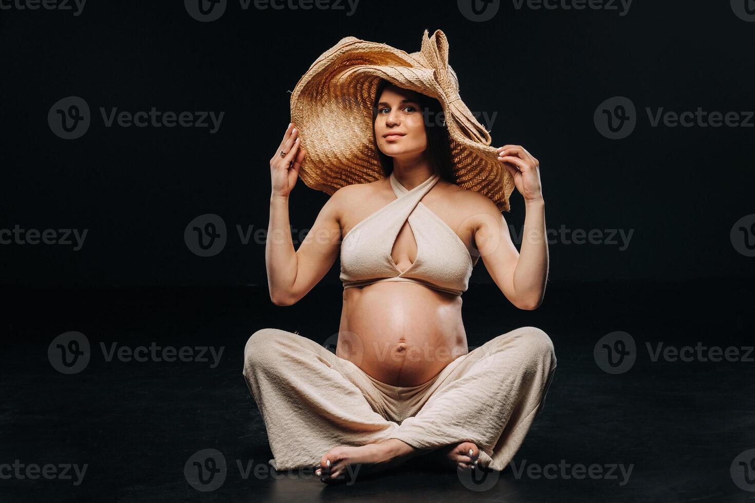 a pregnant woman in a straw hat sits on the floor in beige clothes in a studio on a black background photo