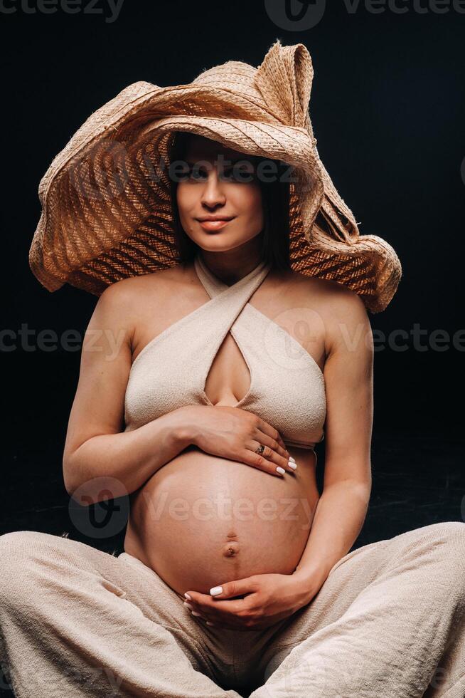a pregnant woman in a straw hat sits on the floor in beige clothes in a studio on a black background photo