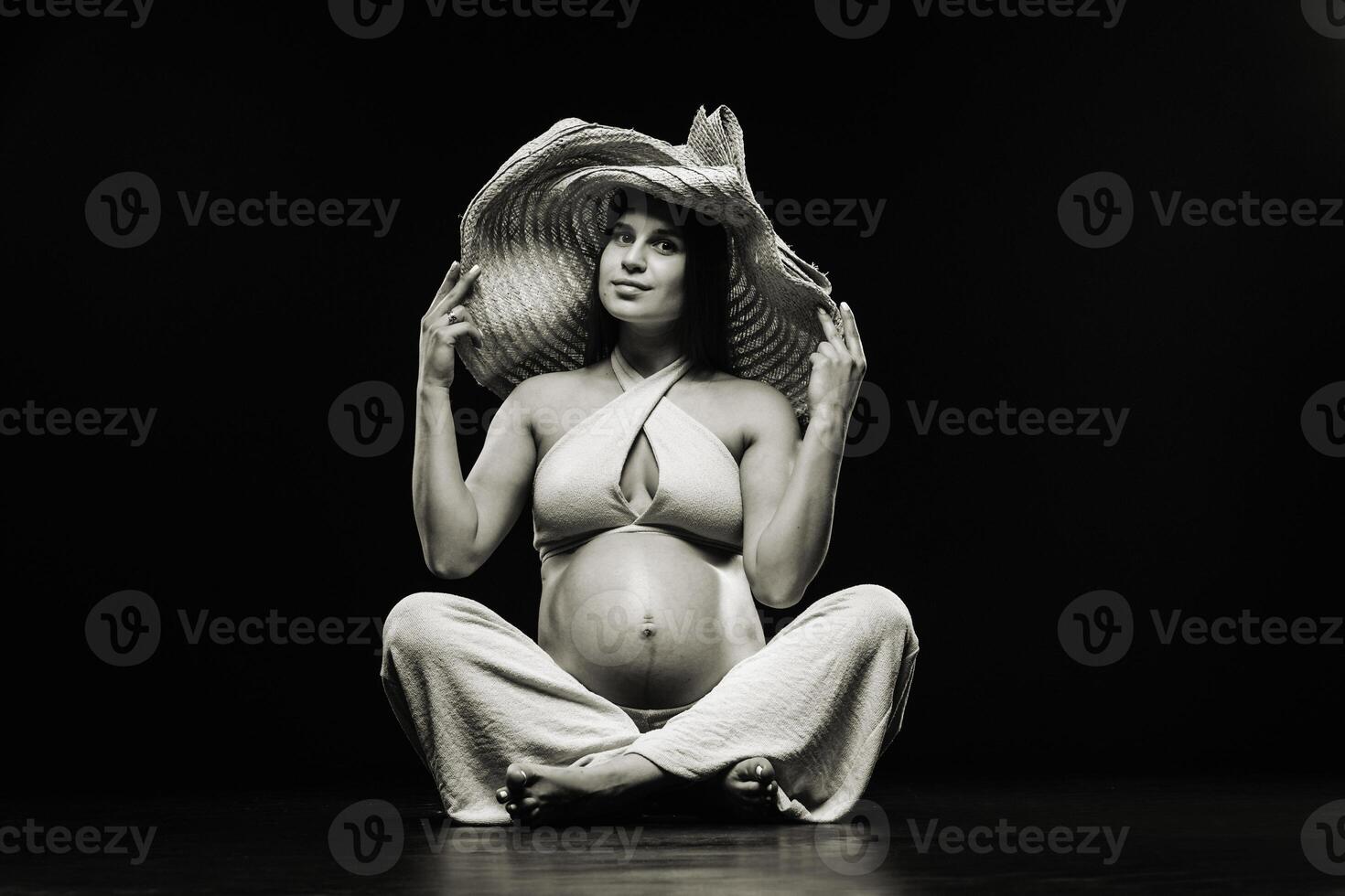a pregnant woman in a straw hat sits on the floor in beige clothes in a studio on a black background photo