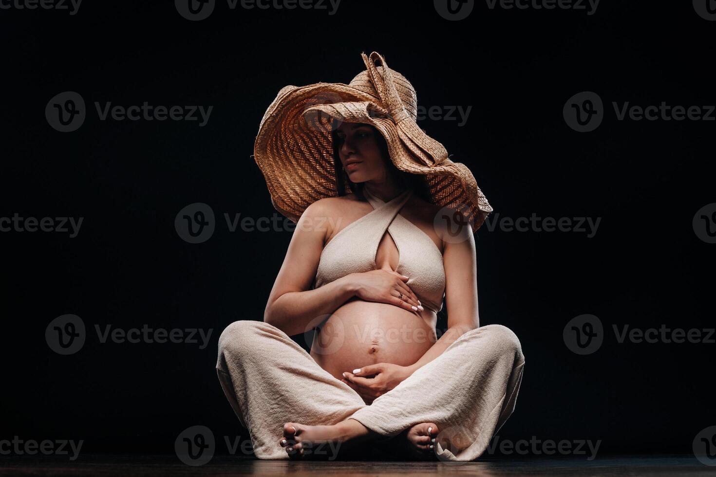 a pregnant woman in a straw hat sits on the floor in beige clothes in a studio on a black background photo