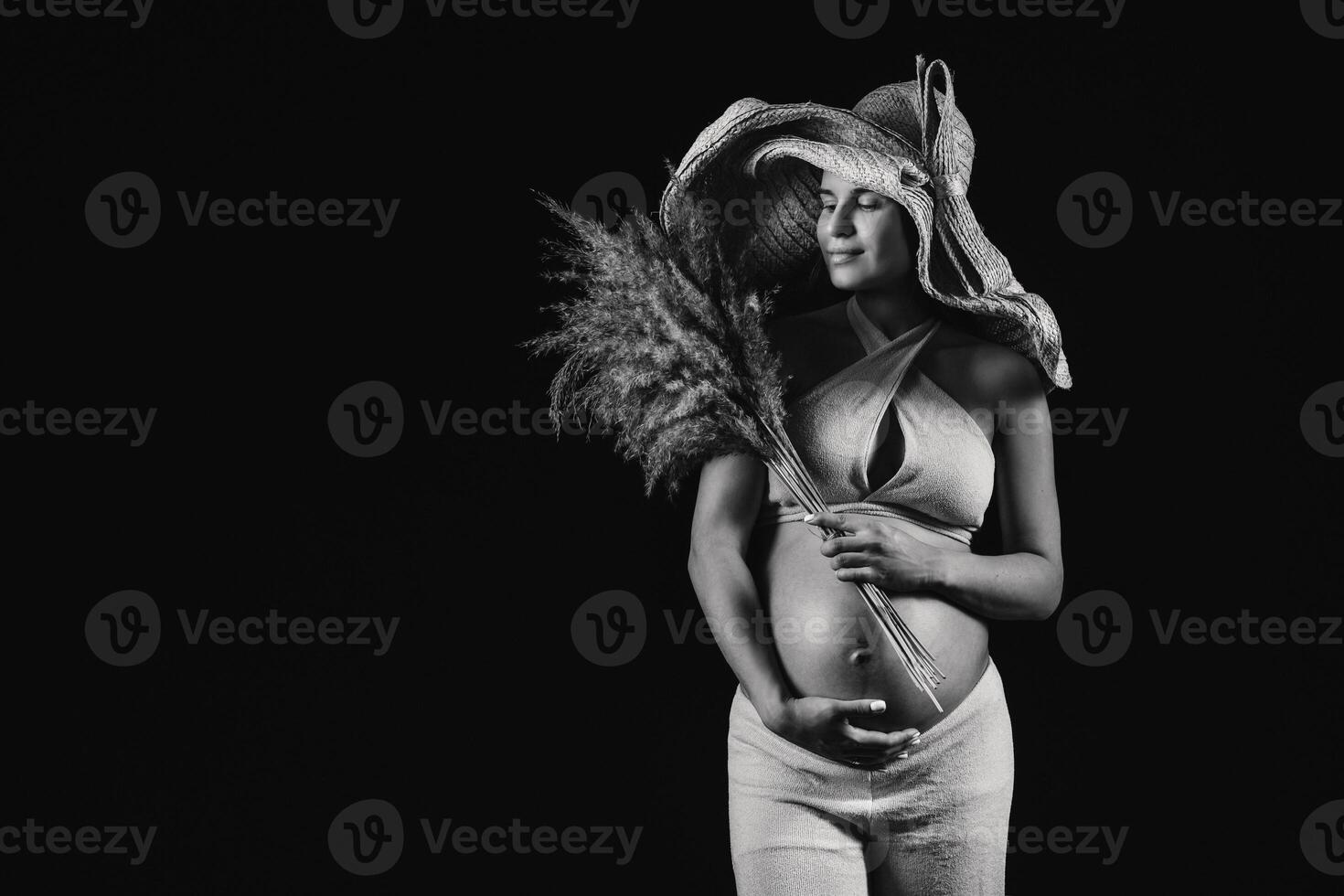 pregnant woman in a straw hat with beige clothes with a bouquet in her hands in the studio on a black background photo
